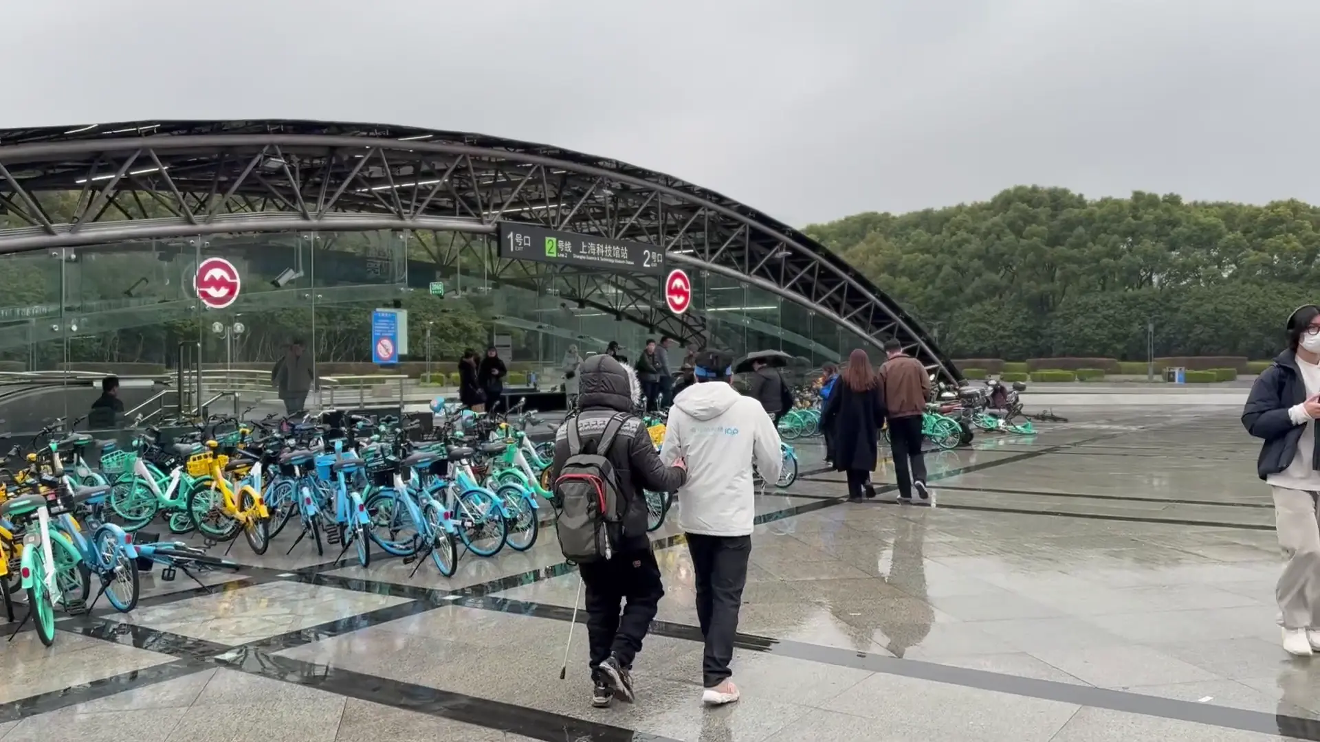Two people walking in the rain in front of a metro station. Many colorful bicycles are in front of the station. One person is guiding the other, who has visual impairments.